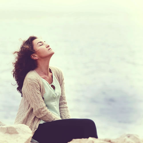A sad woman sitting on the ground near a lake with her eyes closed, face tilted toward the sky to show grieving that can be worked through with a counselor and therapy.