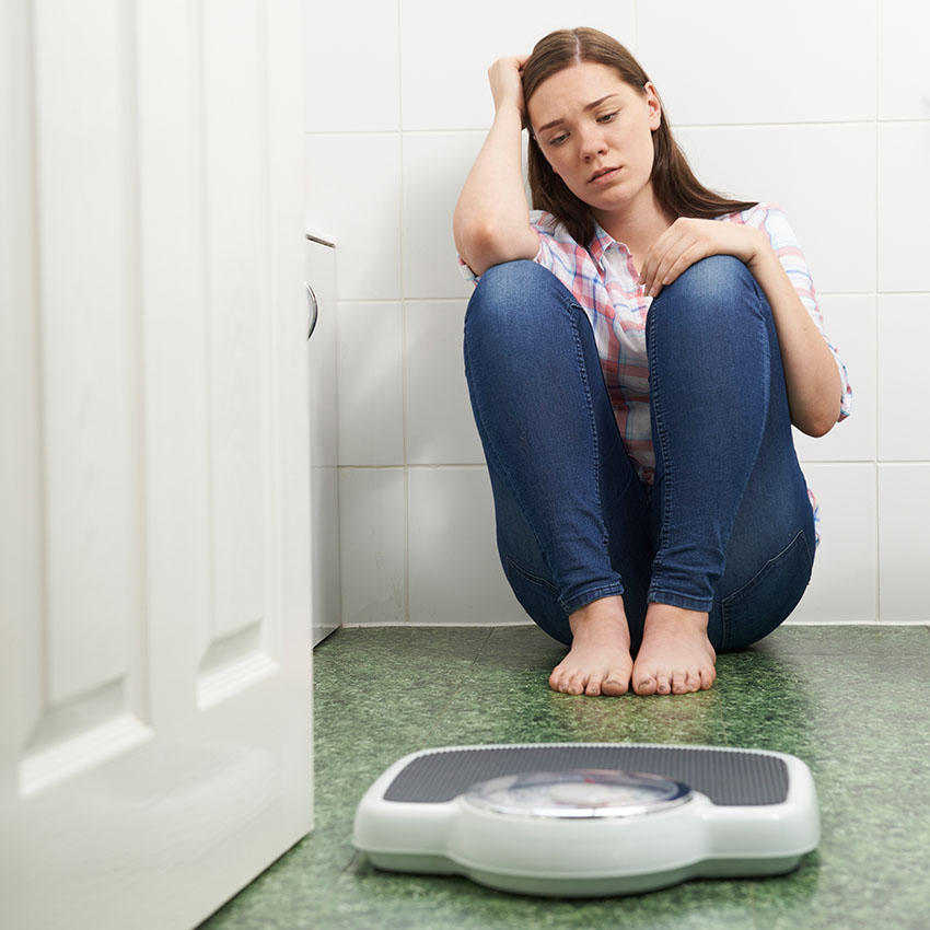 Young, unhappy woman sitting on bathroom floor in front of a scale to show the need negative body image may create that a counselor or therapist can help with.