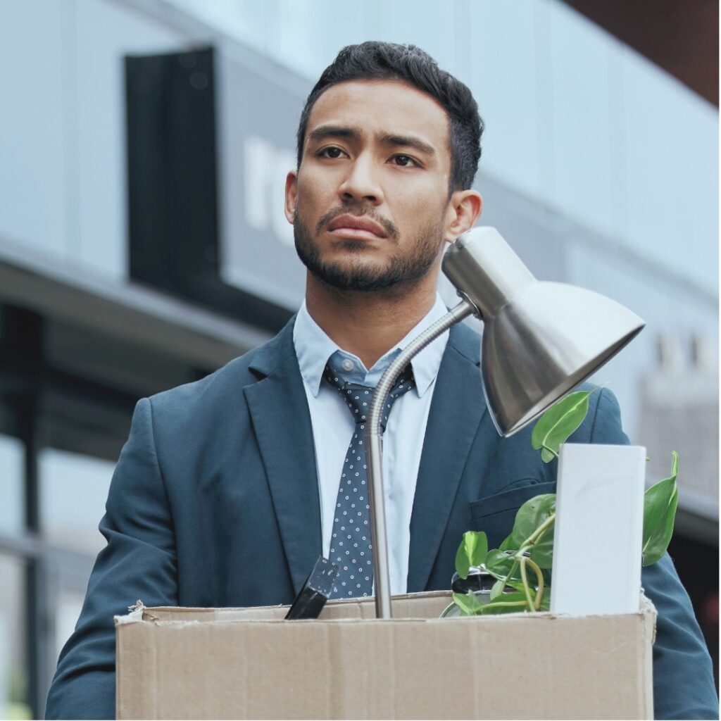 A man with a somber expression, wearing a work suit walking outside a building, and holding a box of possessions including a desk light and plant.