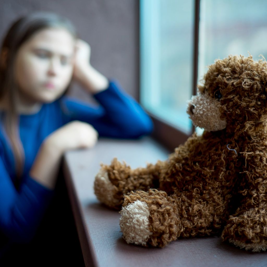 stuffed teddy bear in foreground and sad young girl in background looking out window